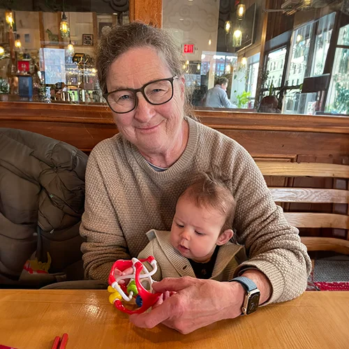 Dr. Andrea Neiley smiling while holding a baby with a colorful toy, sits at a wooden table in a cozy restaurant.